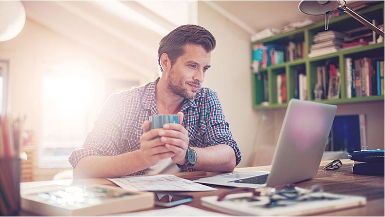 A man is looking at a laptop display while holding a cup in his hands